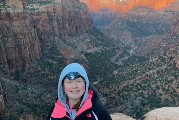 Cameron at dawn standing at the edge of the Canyon Overlook Trail in Zion National Park with a jacket and headlamp on. The sun is just starting to hit the peaks in the background.