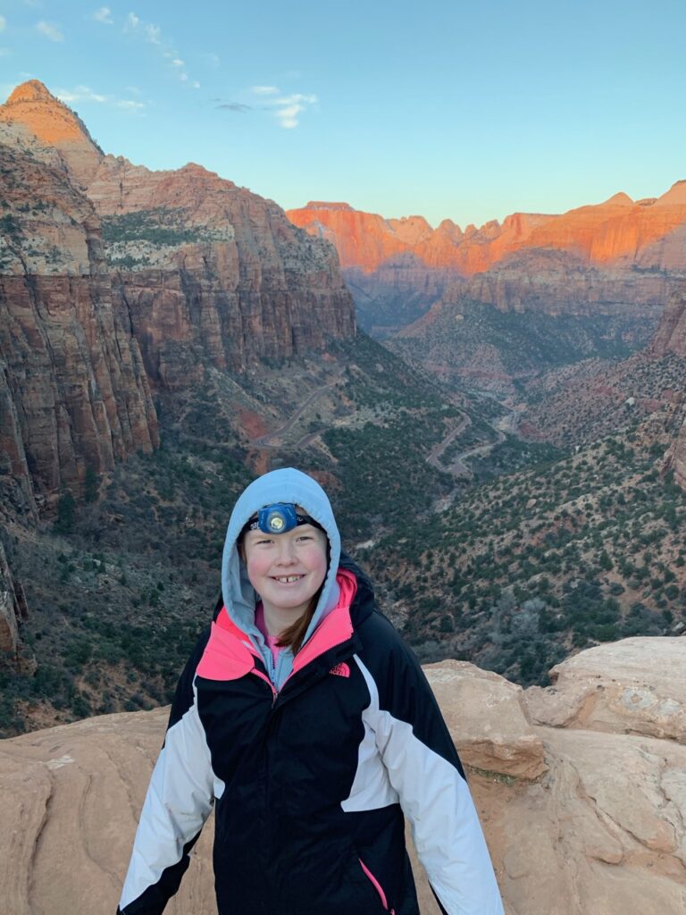 Cameron at dawn standing at the edge of the Canyon Overlook Trail in Zion National Park with a jacket and headlamp on. The sun is just starting to hit the peaks in the background.