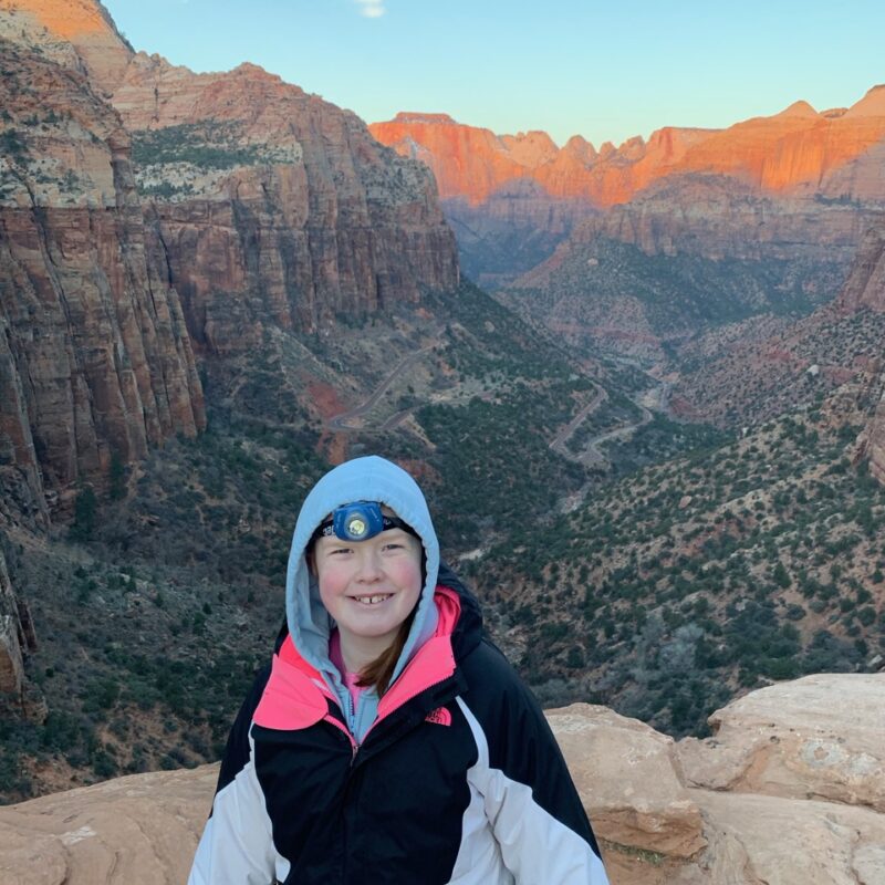 Cameron at dawn standing at the edge of the Canyon Overlook Trail in Zion National Park with a jacket and headlamp on. The sun is just starting to hit the peaks in the background.