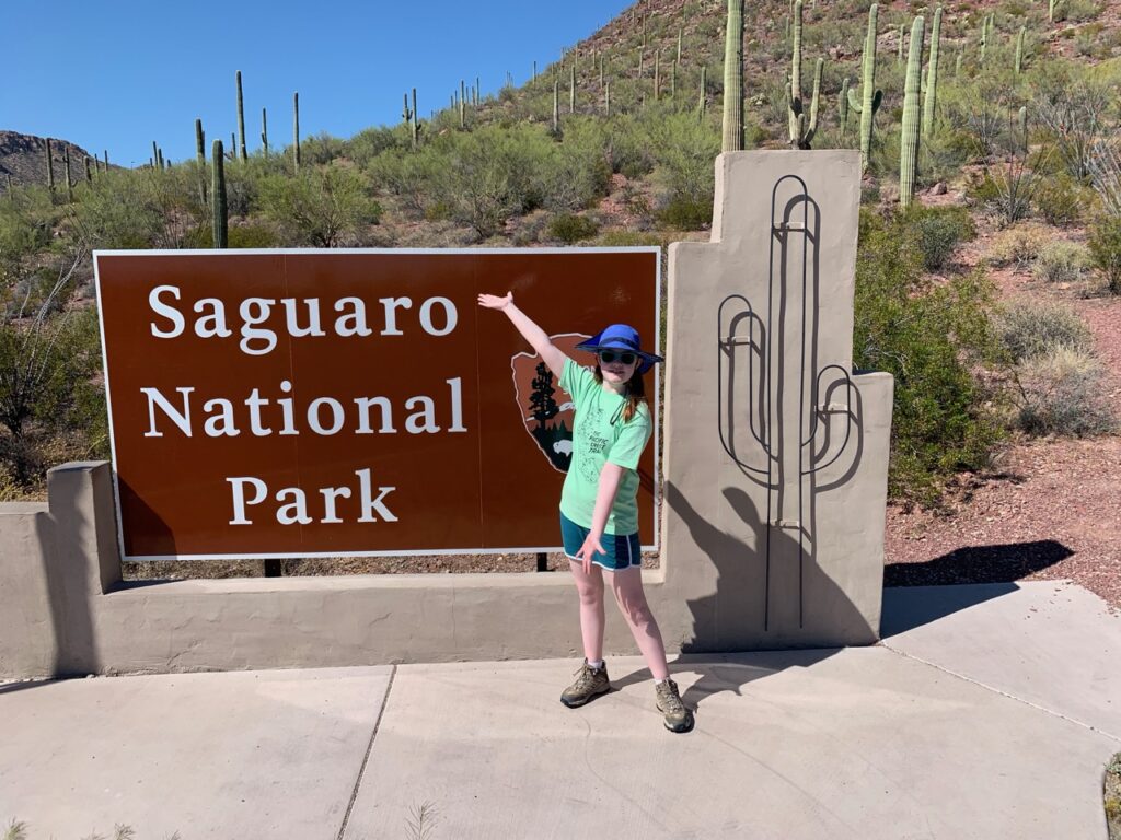 Cameron standing in front of the Saguaro National Park sign in shorts, hat and sunglasses. 