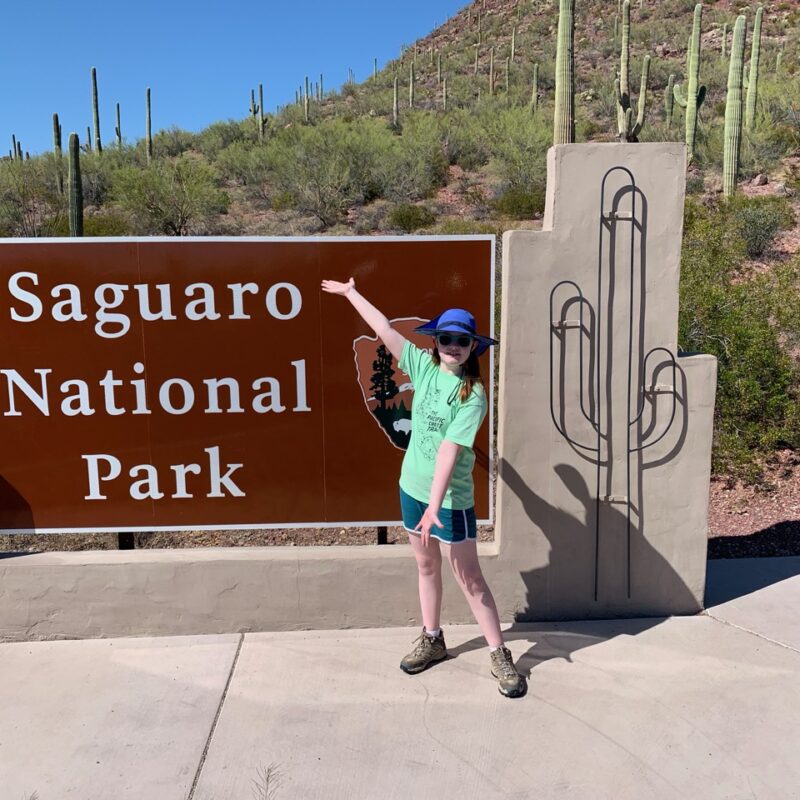 Cameron standing in front of the Saguaro National Park sign in shorts, hat and sunglasses.