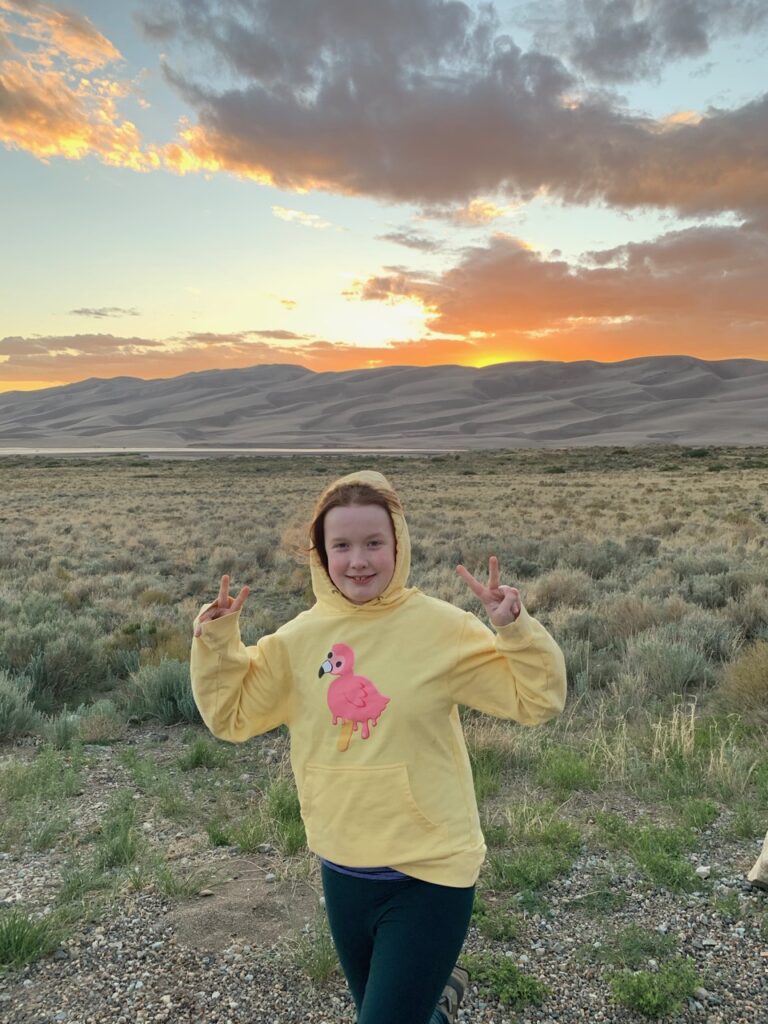 Cameron standing on the grasslands in front of the sand dunes - the sky is full of clouds and color at sunset. 