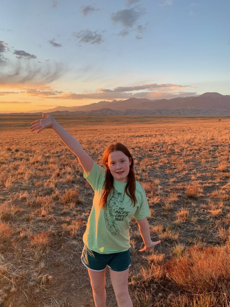 Cameron standing in front of the sand dunes and mountains, the sky is full of color and clouds at sunset. The warm list is cast over her making her glow red.