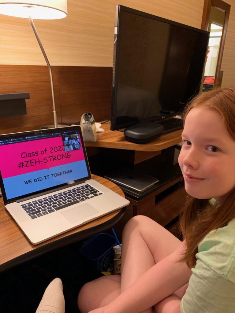 Cameron sitting at the desk in our hotel room, with her mac laptop open for her remote Zoom 5th grade graduation. 