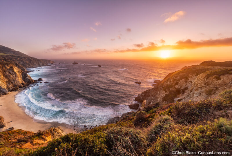 Sunset over the Pacific Ocean near Bixby Bridge on California's central coast. A breathtaking scene with clouds above the horizon, creating a vibrant sunset, while a sun star appears as the sun dips below. The image captures the rugged beauty of Big Sur, featuring steep cliffs, mountains in the distance, and a half-moon bay with deep blue waters. This composition reflects the striking and serene landscapes that define the central California coastline.