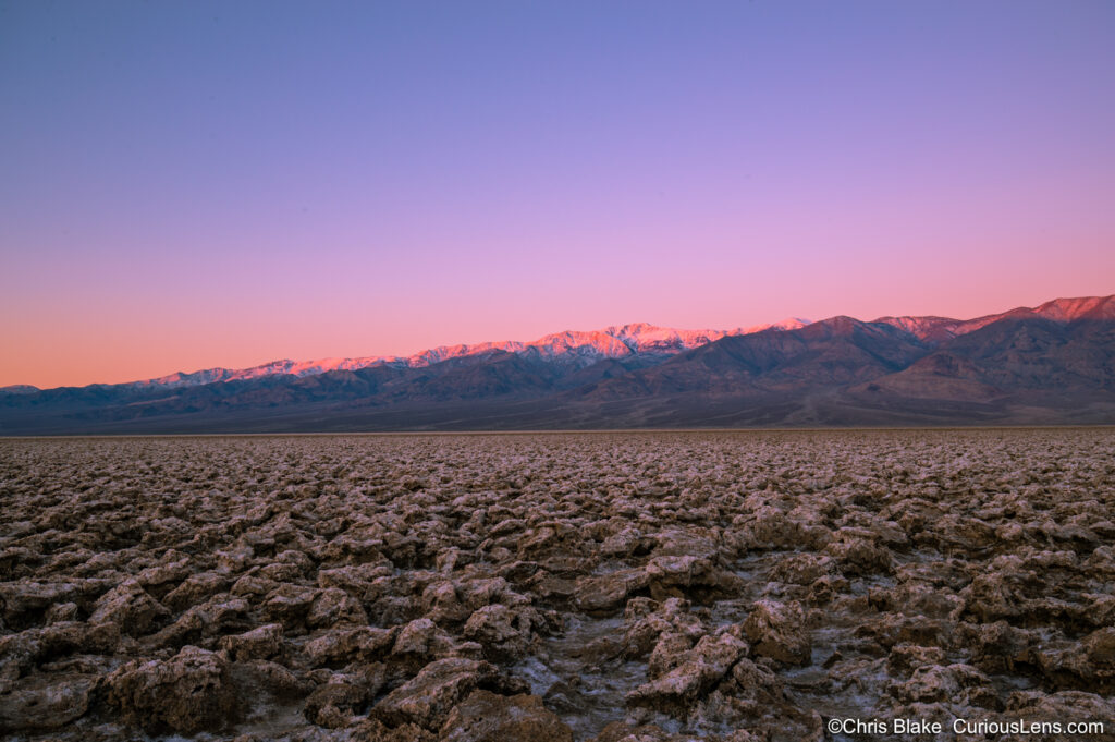 Panoramic view of the Devil's Golf Course in Death Valley National Park at dawn. The sun rises over the Panamint Range, bathing the salt pan in a warm golden light. The scene is tranquil and eerily quiet, capturing the otherworldly beauty of this unique desert landscape.