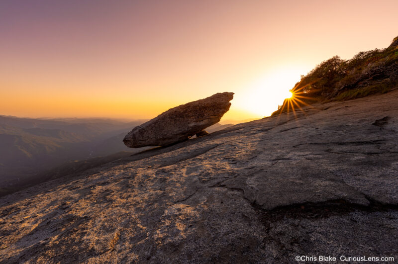 A scenic view from the Hanging Rock Trail in a sequoia forest, showing the top of a granite mountain. The foreground features the iconic Hanging Rock, with the sun creating a starburst effect as it sets behind the horizon. The sky glows with vibrant shades of yellow and red, indicating the onset of sunset. The last rays of sunlight cast a warm glow on the rock formations. In the distance, there's a view of Moro Rock, and the valley below is filled with smoke, adding a sense of drama to the scene.