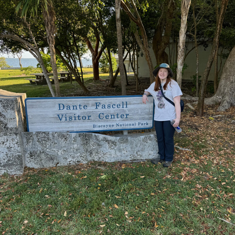 Cameron standing in front of the sign for Dante Fascell Visitors Center in Biscayne National Park. She is wearing a hat and has a huge smile on her face.