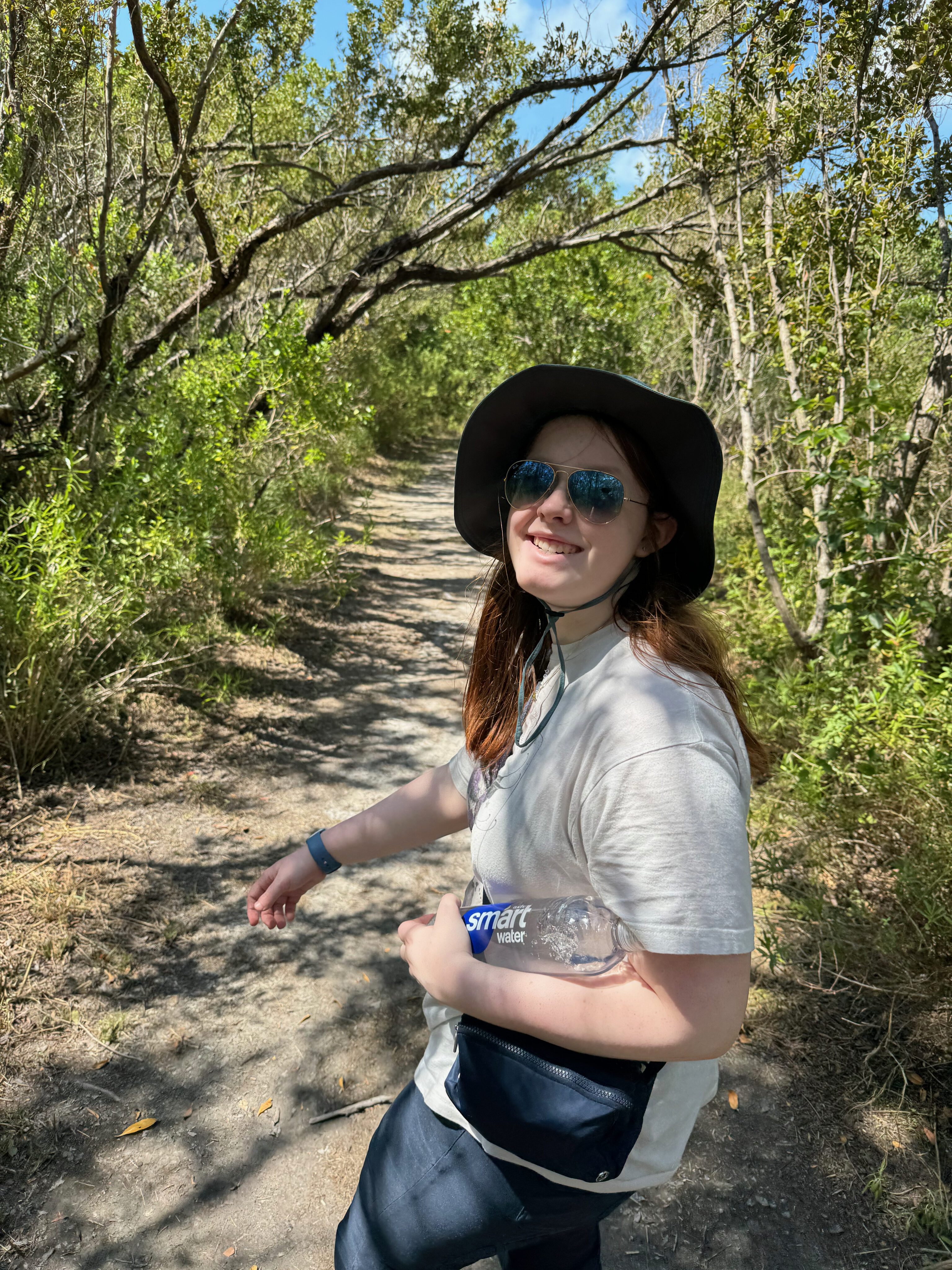 Cameron wearing sunglasses and a hat smiles as she hikes through the deep words on Boca Chita Key.