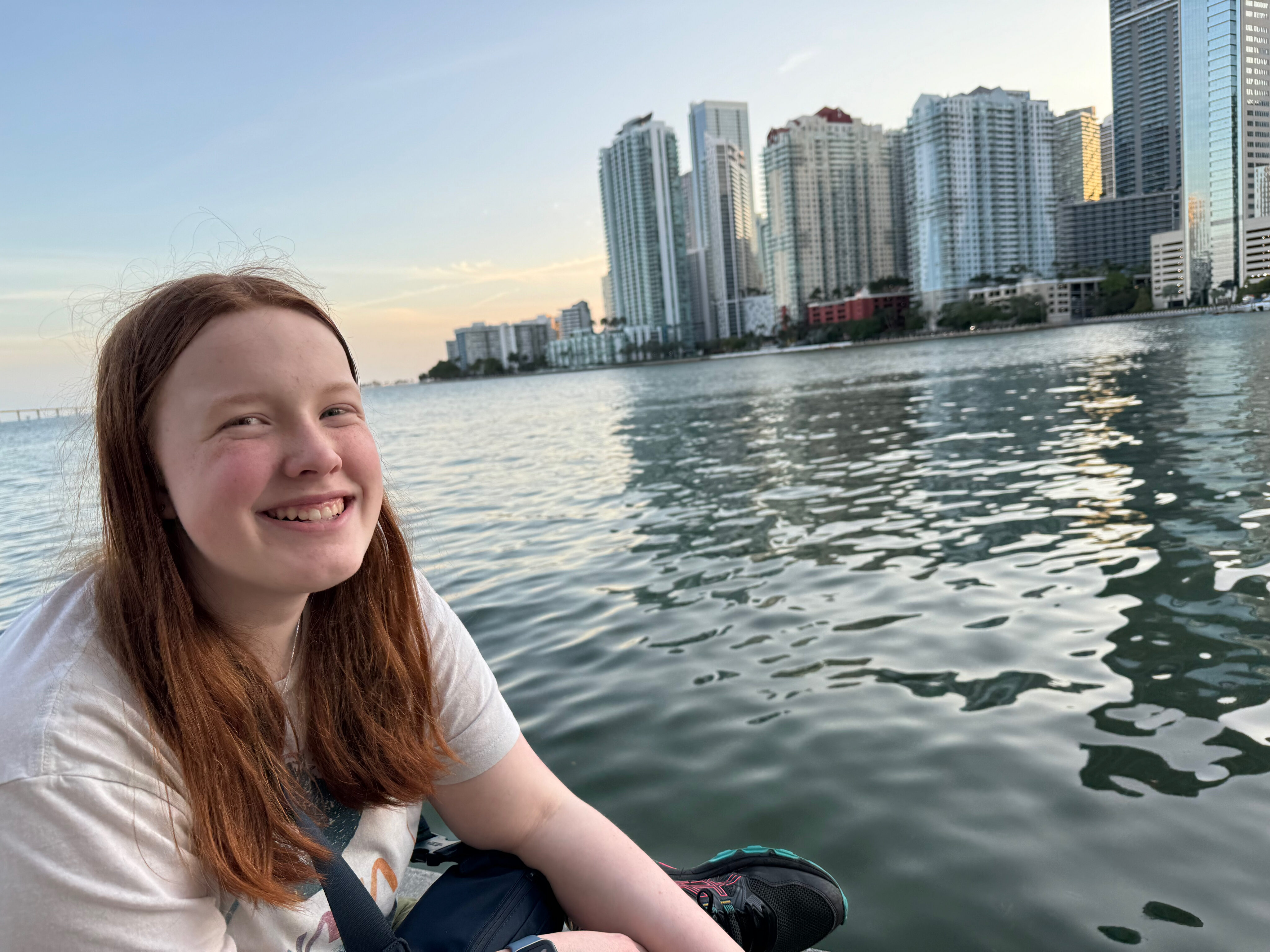 Cameron sitting on the wall, with a huge smile on her face on Claughton Island. The bay and Miami skyline can be seen behind her.