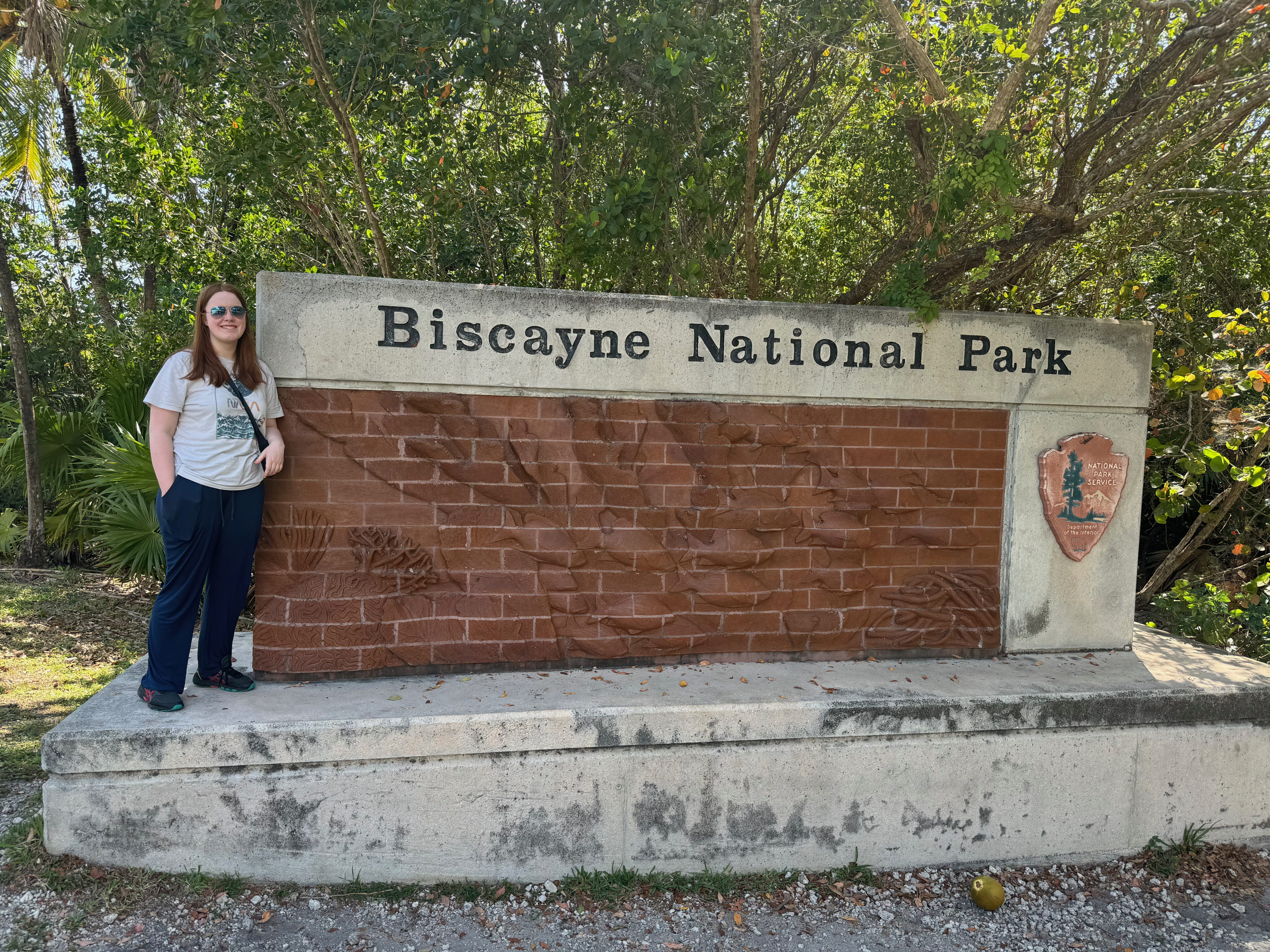 Cameron wearing pants, a t-shirt and sunglasses standing next to the part sign for Biscayne National Park. 