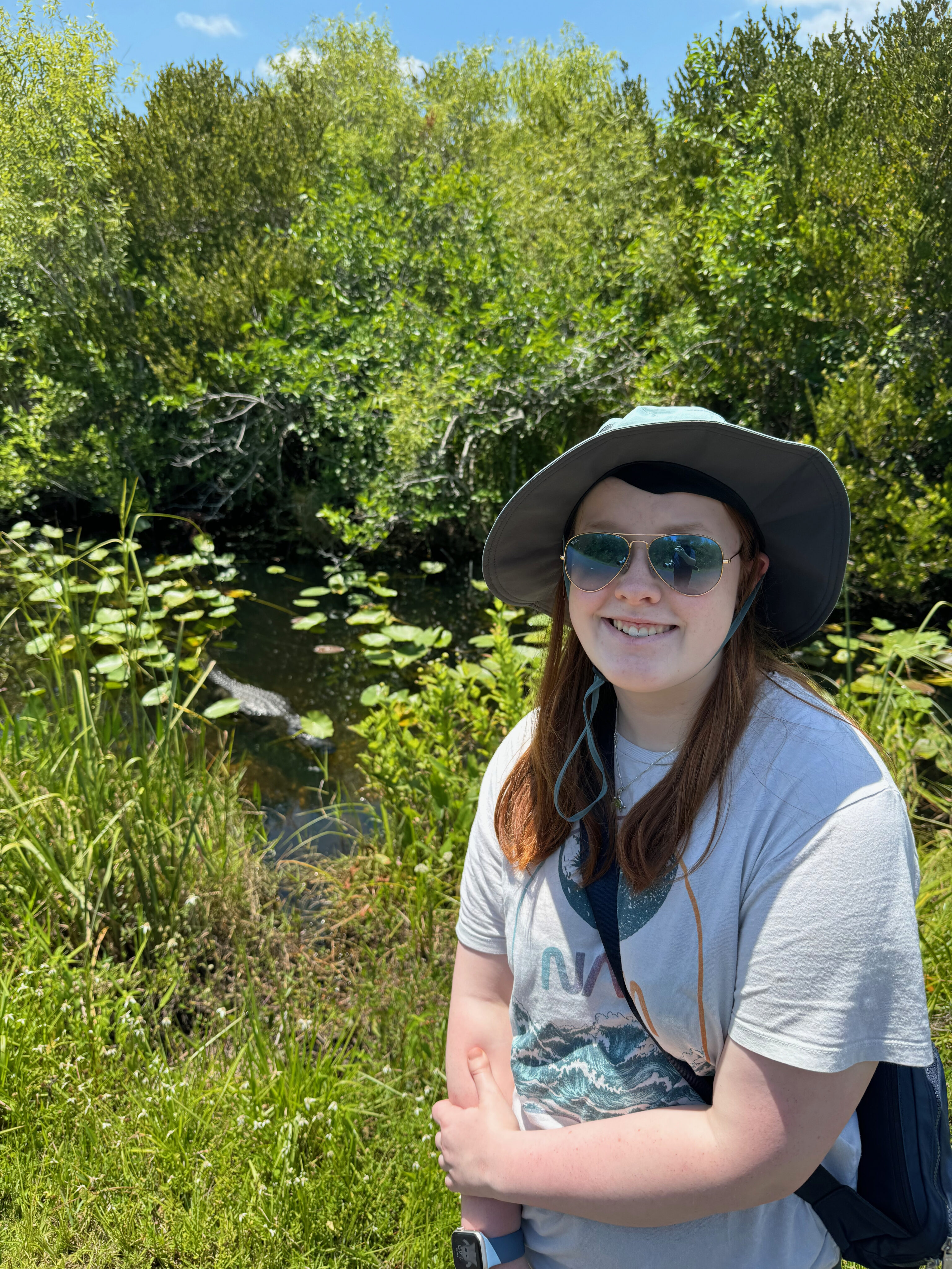 Cameron wearing a hate and sunglasses stops for a photo on the Shark Valley Loop Trail. Behind her there is an alligator in the water. 