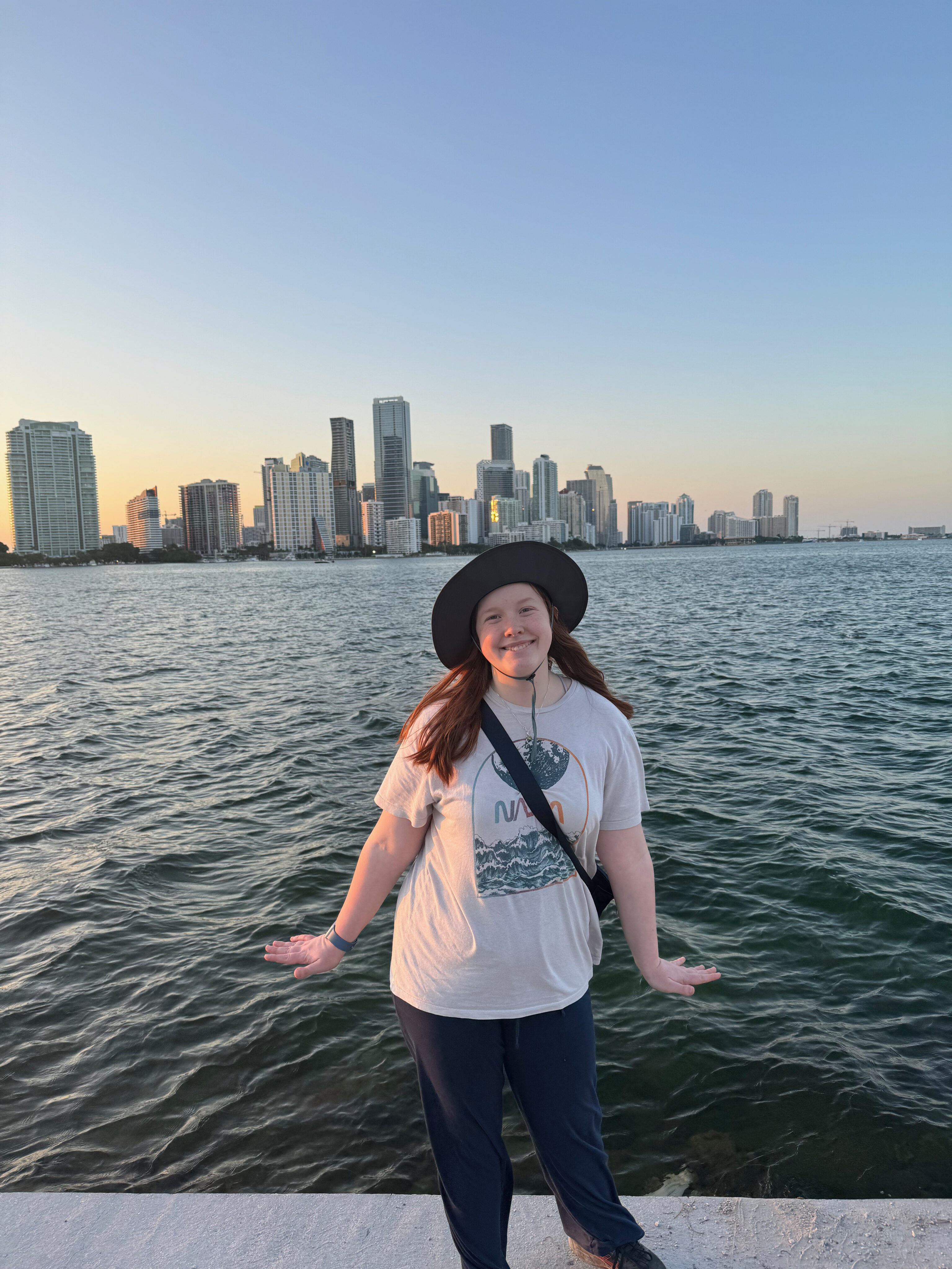 Cameron standing on the stone seawall off the Rickenbacker cswy with the Miami skyline behind her. The sun had just set and the sky is turning yellow.