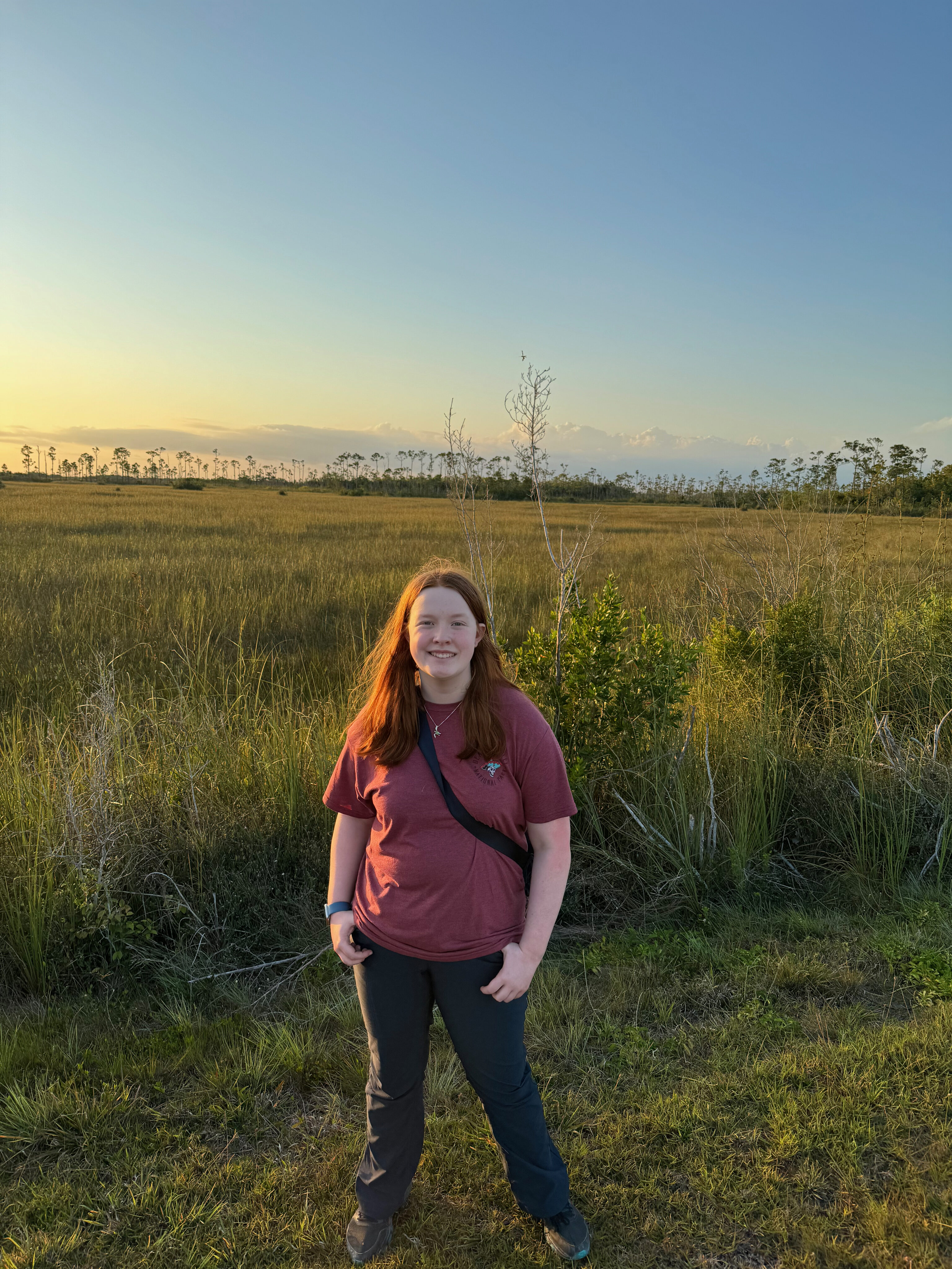 Cameron standing near a tall field in the everglades at sunset. A few clouds on the horizon are just starting to turn colors at sunset. 