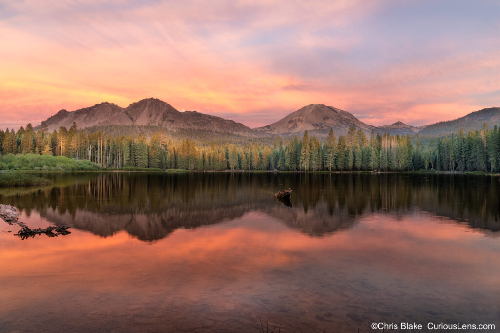 Scenic view of Manzanita Lake at sunset with a backdrop of Lassen Peak, showcasing vibrant clouds and the last light illuminating the mountain and treetops. The tranquil lake reflects its volcanic origins, with an easy walking trail encircling the water, capturing the natural beauty of the landscape.