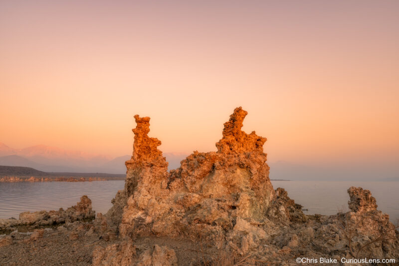 An early morning scene at Mono Lake in the Eastern Sierra. The photo captures the sun as it crests the horizon, bathing the landscape in a warm, golden light. The unique tufas in the foreground glow with a vivid orange color, while the snow-capped peaks of the eastern Sierra mountains in the background catch the first rays of sunlight. The entire sky is illuminated with a gentle yellow hue, contributing to the serene and tranquil atmosphere."