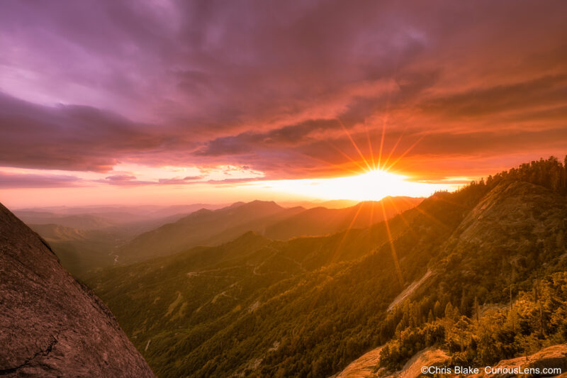 View from Moro Rock in Sequoia National Park, 6,725 feet above sea level. The sunset creates a sun star as it dips below the mountains, casting a warm glow on clouds. Thick haze and smoke from nearby forest fires are visible in the San Joaquin Valley below. The scene is reached by climbing 350 granite steps to the summit.