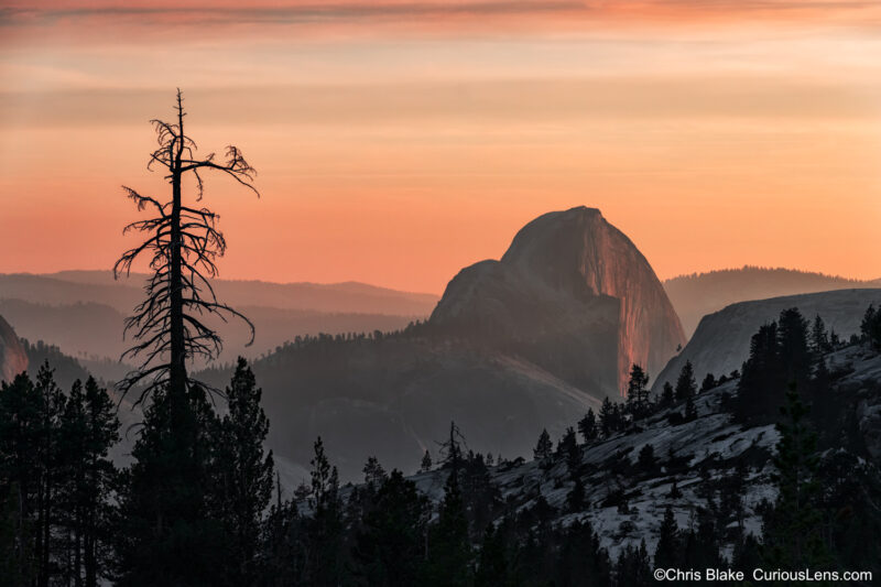 View of Clouds Rest and Half Dome from Olmsted Point in Yosemite National Park at sunset. The sky glows with vibrant colors, and Half Dome has a striking red hue. Smoke from the "Camp Fire" contributes to the haze, creating an atmospheric effect. This stunning scene is a reminder of the park's beauty and the broader impact of California's wildfires.