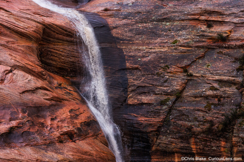 A post-rain landscape illuminated by a golden sunset, where streams of melted snow and rainwater flow down steep cliffs, filling dry riverbeds. Spontaneous waterfalls tumble over fiery red rocks, creating a beautiful and contrasting scene of gentle water against rugged terrain.