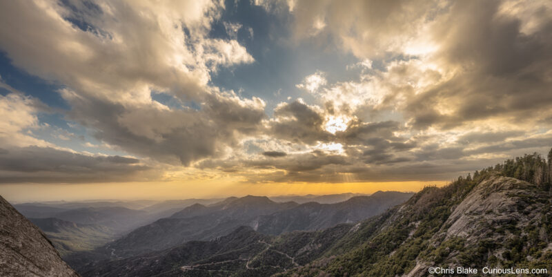 A panoramic view from Moro Rock in Sequoia National Park, with dark storm clouds covering the sky. Rays of sunlight break through the clouds, casting light on the foothills and the San Joaquin Valley. This photo was taken about an hour before sunset.