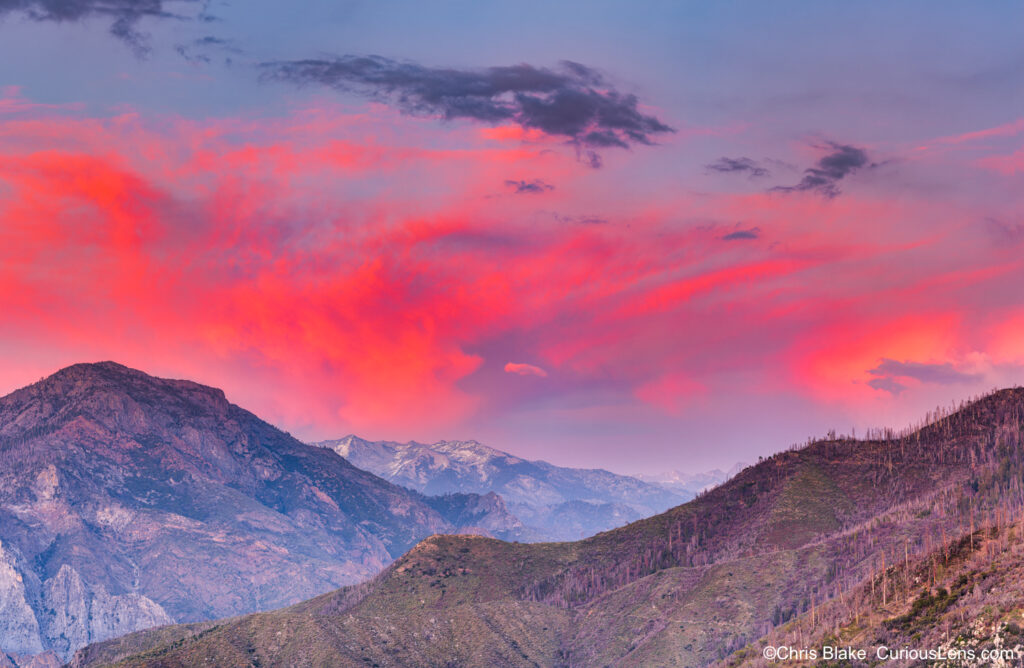 View of the Great Western Divide in the Sierra Nevada Mountain Range at sunset. The scene shows partially snow-covered peaks in the distance, with the vibrant colors of a spring evening sky. Photo taken from an unmarked bend on Hume Lake Road, near Kings Canyon National Park.