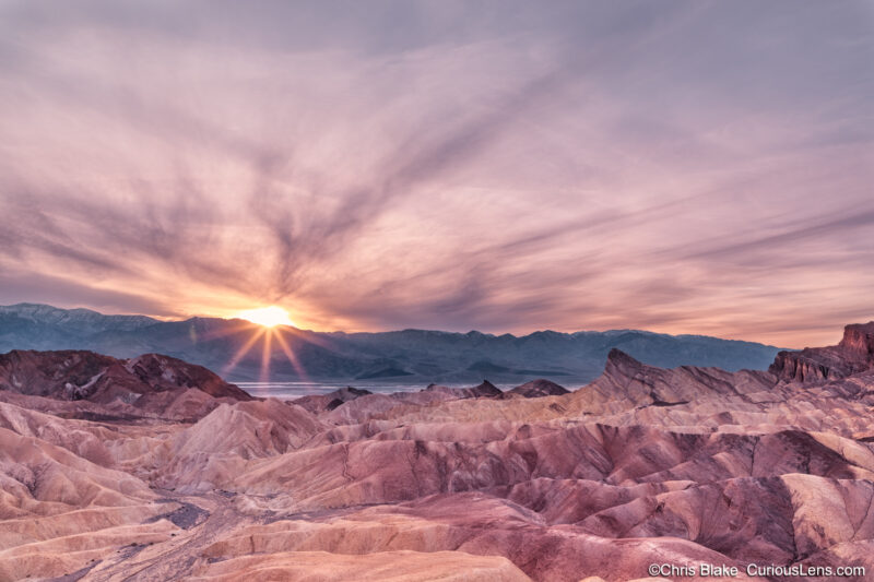 Zabriskie Point in Death Valley offers stunning views of the Amargosa Range and Panamint Mountains. The foreground features the iconic Manly Beacon with the expansive Death Valley Basin stretching into the distance. This time-blended image combines the vivid colors of sunset and the serene glow of the blue hour, capturing the vibrant hues and breathtaking landscape of this popular location.