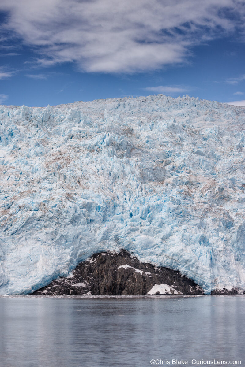 Aialik Glacier in Kenai Fjords National Park with mirror-like waters, icy facade, rugged triangular rock outcropping, and deep blue sky with wispy clouds.