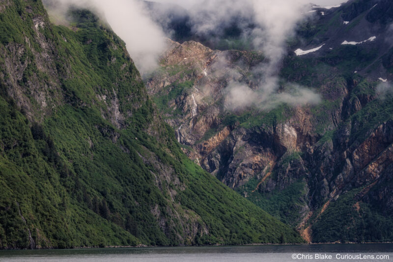 Aialik Bay in Kenai Fjords with towering cliffs, lush and rugged rock faces, and a cascading waterfall carving a valley into the mountainside. Late afternoon light and clear view after clouds break.