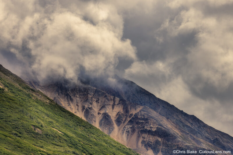 Wrangell mountainside with vibrant green grass, ferns, and lush trees in dappled sunlight transitioning to dark rock. Dramatic storm clouds cascade down, transforming the landscape.