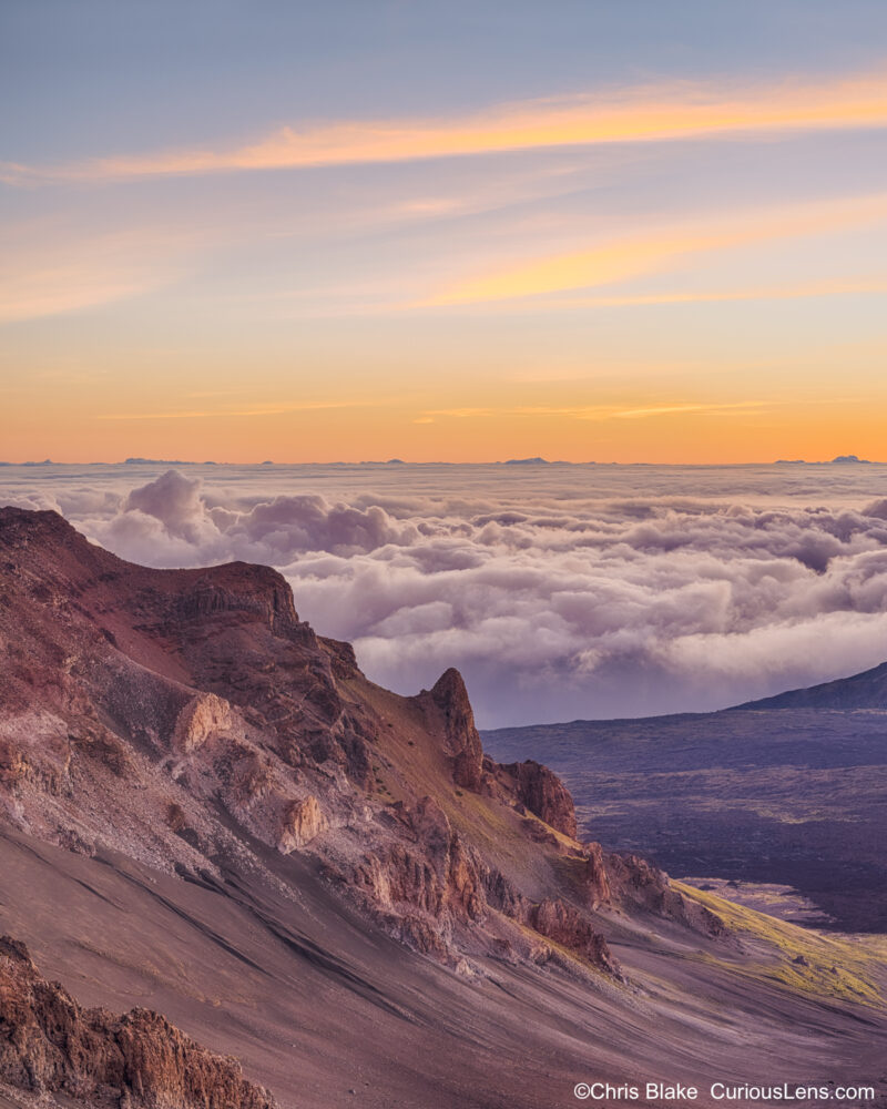 unrise at East Maui Volcano in Haleakalā National Park, showcasing the contrast between soft clouds and warm pre-dawn light against the jagged rocks. Captured above the cloud inversion at over 10,000 feet, this image offers a surreal view of the cinder-cone volcano illuminated by the rising sun.