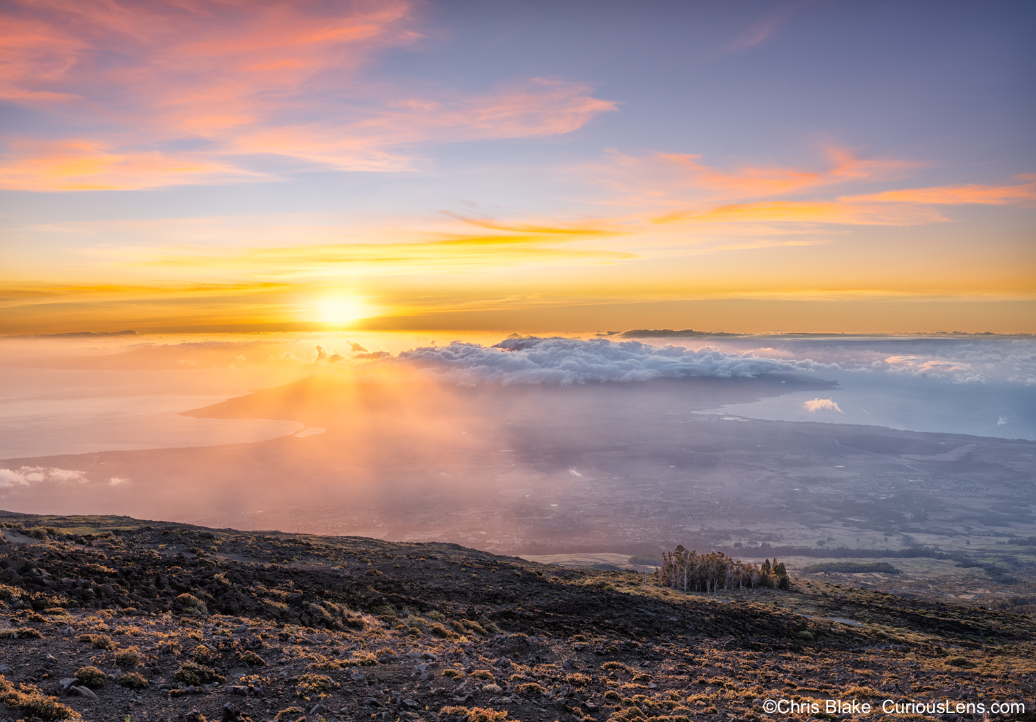 Sunset time-blend photo taken from Haleakalā Summit in Maui, showcasing warm yellow-orange hues washing over clouds with ocean visible below. Trees below illuminated by last light.