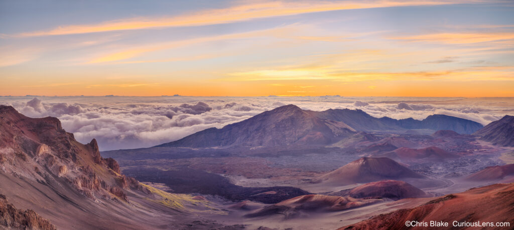 Panoramic sunrise over East Maui Volcano in Haleakalā National Park, with warm blue hour light illuminating the crater below. The sky glows yellow as the sun rises through the clouds, revealing cinder cone volcanoes.