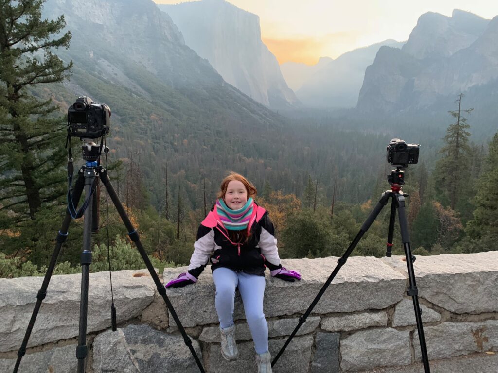 Cameron sitting on the stone wall at Tunnel View in Yosemite National Park. Photo is taken at sunrise with a red sky, Half Dome and El Capitan in the background.