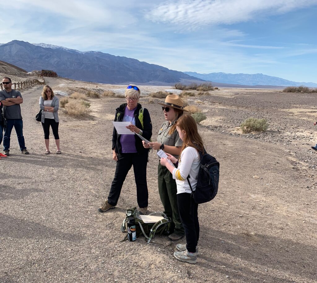 Cameron in death valley, reading to a group. With Park rangers and a blue sky with clouds overhead. 