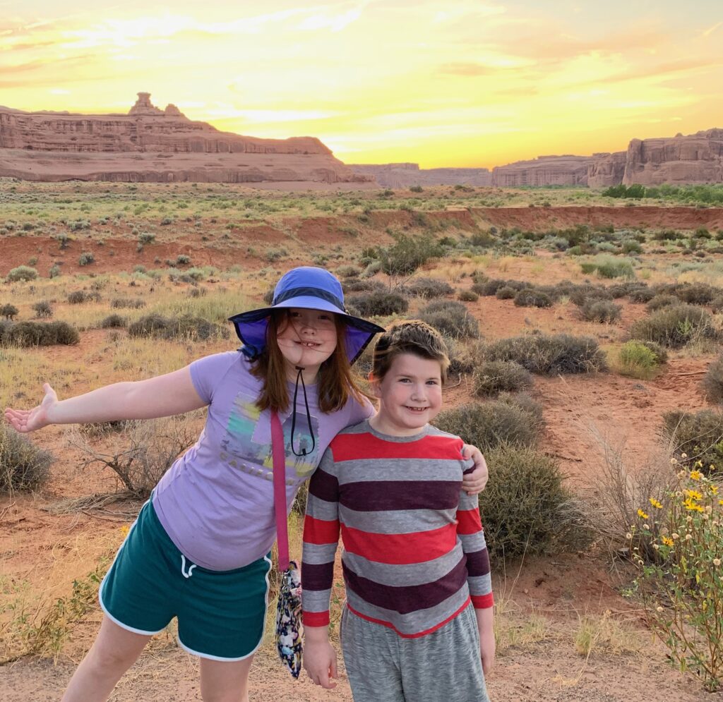 Cameron and Collin standing in the desert at Sunset in Arches National Park. The sky is bright yellow and full of color at sunset.