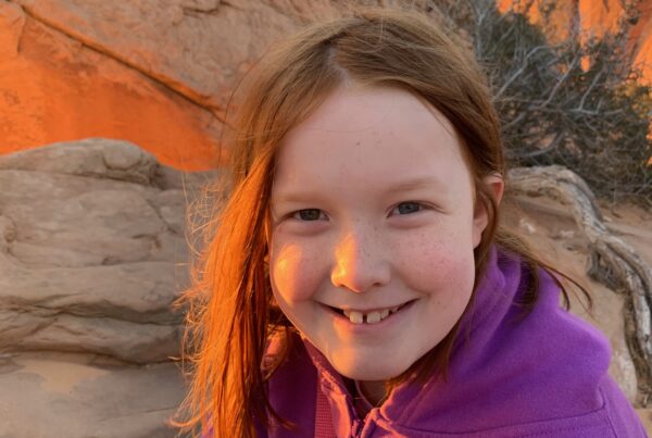 Cameron sitting at Mesa Arch at sunrise, with the warm red light washing across her face.