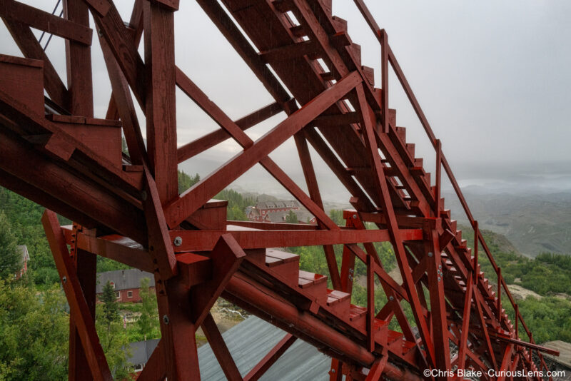 View from the ore processing building in historic Kennecott, Alaska. Vibrant red ore chute contrasts with dark gray skies and the green valley below, where a glacier once stood.