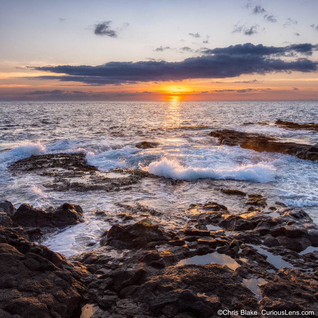 Sunbeam breaking through clouds over the horizon, illuminating a small wave in front of a brimming lava tube. The unique photograph captures the interplay of light across the water in a serene and distinctive scene.