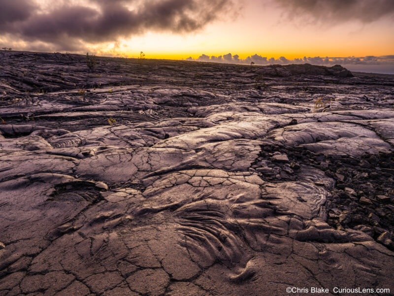 Sunrise over lava fields, where early morning light enhances the rugged textures and contrast of the foreground. The sky glows yellow, with clouds adding striking contrast against the brightening horizon.