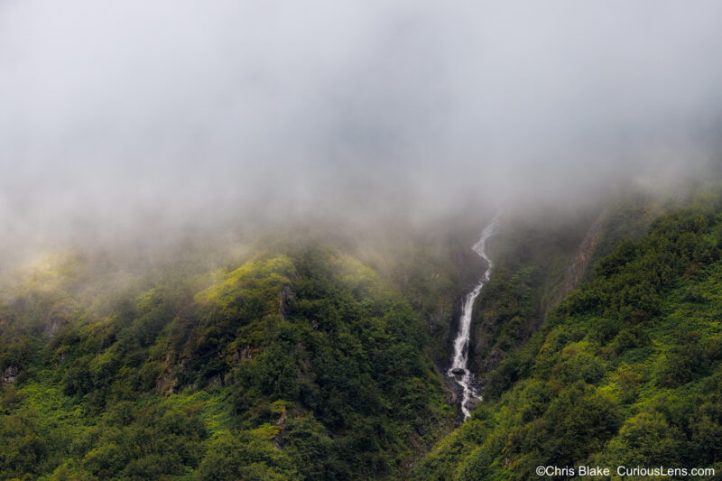 Exit Glacier with sun piercing through clouds, illuminating fog-covered mountains and vibrant greens. Waterfall on the far right anchors the photograph.