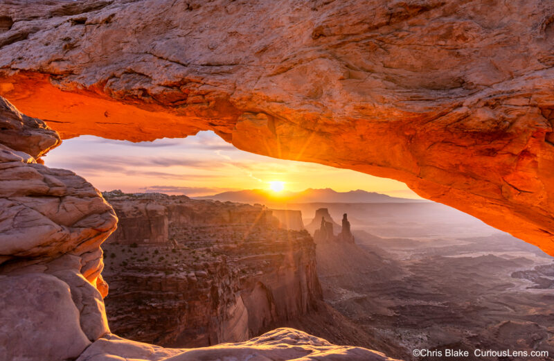 Mesa Arch at Canyonlands National Park in the Island in the Sky district, overlooking a canyon with views of Monster Tower, Washer Woman Arch, Airport Tower, and the La Sal Mountains. The photo captures a unique angle from the far right, featuring a sunburst through the arch, adding a dramatic effect to the landscape scene.