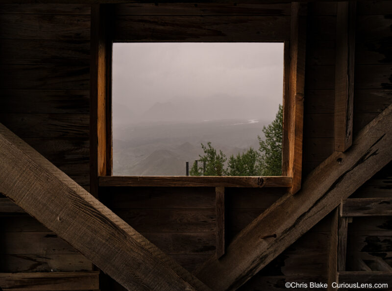 Explore Kennecott, Alaska's historic town with iconic mills and mines. Preserved by the National Park Service, it reflects its 1920s copper production heyday. Tour the colossal ore processing building, with its awe-inspiring size and century-old wooden frames. On a rainy August afternoon, discover an old window casting a warm glow, with the valley below shrouded in clouds and rain.