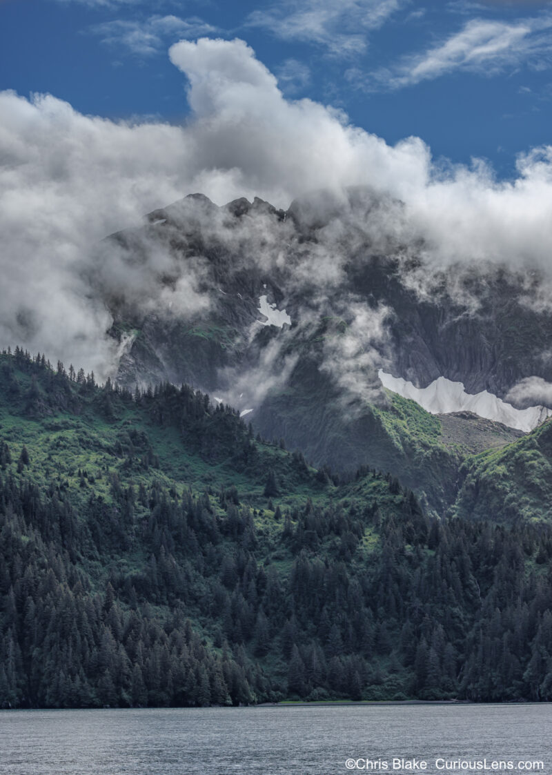 Kenai Fjords National Park with tranquil, mirror-like waters, lush green fir trees, rugged mountain peaks, glaciers, low-hanging clouds, and a deep blue sky.