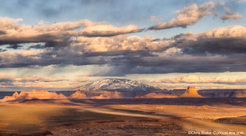 A stunning composite image of Navajo Mountain, located near Page, Arizona, standing at 10,388 feet and covered in snow. The photo is taken from a high vantage point atop a neighboring mountain, with the mountain centered in the background. In the foreground, striking rock formations add texture, while the sky features dramatic clouds dumping rain and snow in the distance. The sunset creates a dynamic interplay of light and shadow, contributing to the surreal and captivating quality of the scene.