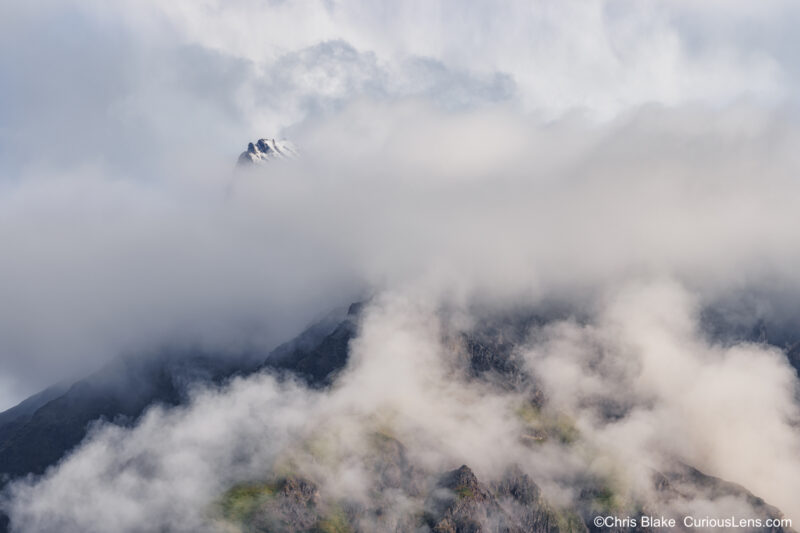 Regal Mountain in Wrangell-Saint Elias National Park. Eroded stratovolcano at 13,999 feet, partially shrouded in glaciers. Peak illuminated by sunlight, with clouds parting to reveal dark rocks and vibrant green grass below."