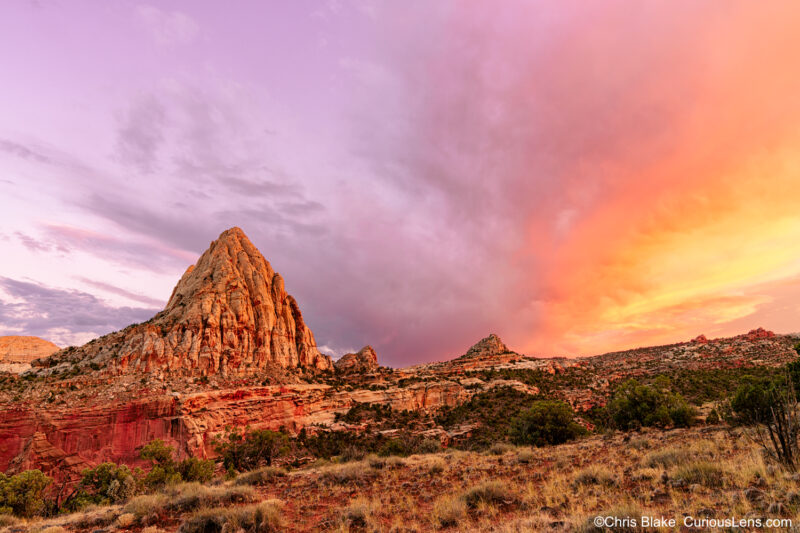 A vibrant sunset at Capitol Reef National Park with Pectols Pyramid in the background. The sky glows red as it sets behind the iconic 6,211-foot-tall landmark. The photo was captured after a rainy day with partially washed-out trails, offering a unique and dramatic composition.