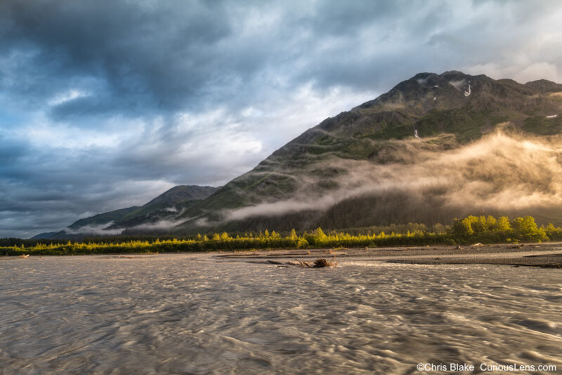 Resurrection River winding through Kenai Peninsula to Resurrection Bay, near Seward, Alaska. Ancient bridge, storm clouds, low-lying clouds, and sunlit trees create a fleeting, magical scene.