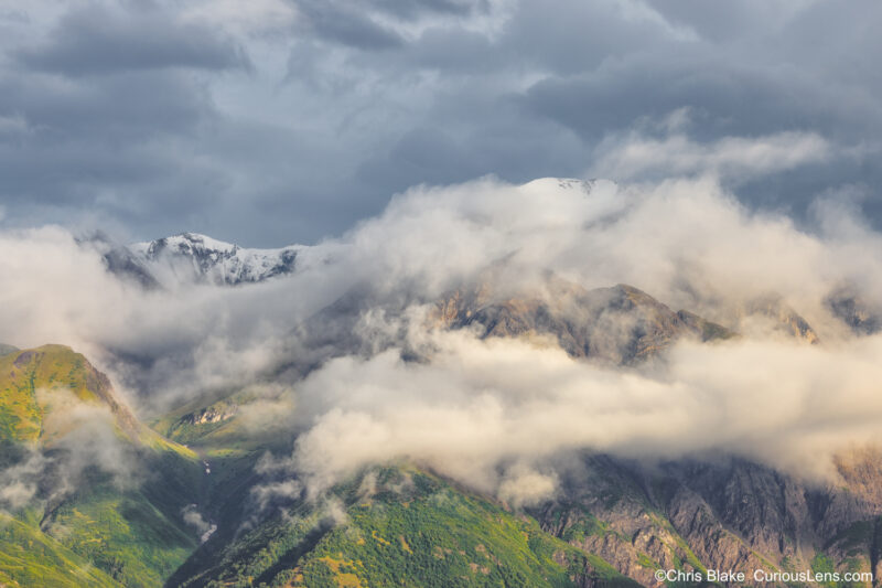 Fireweed Mountain Range in Alaska, showcasing lush green valleys, snowcapped peaks, and dramatic clouds. Morning light illuminates the landscape, highlighting the transition from verdant valleys to rocky terrain. A stunning example of Alaska's natural contrasts and beauty.