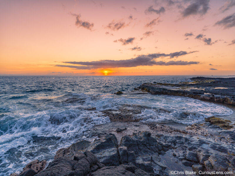 Sunset at Kohanaiki Beach Park (Pine Trees Beach) near Kailua-Kona, featuring serene lava tubes with water gently washing over them, framed by preserved trees and a warm, colorful sky. The peaceful shoreline blends sandy beaches with lava rock, capturing the tranquil beauty and solitude of the Hawaii coastline.