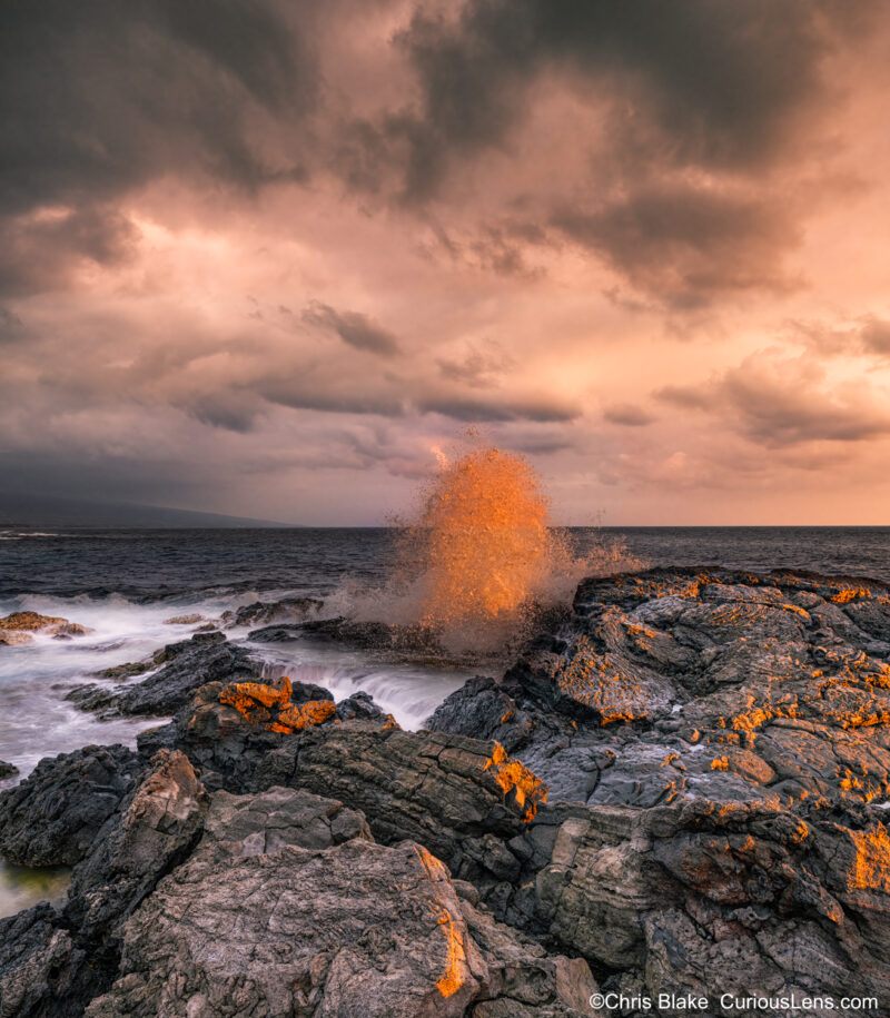 Sunset over a Hawaiian beach with rocks warmly lit by the sun peeking through storm clouds. The water glows red as it crashes, while spray frozen mid-air adds contrast against the stormy sky.